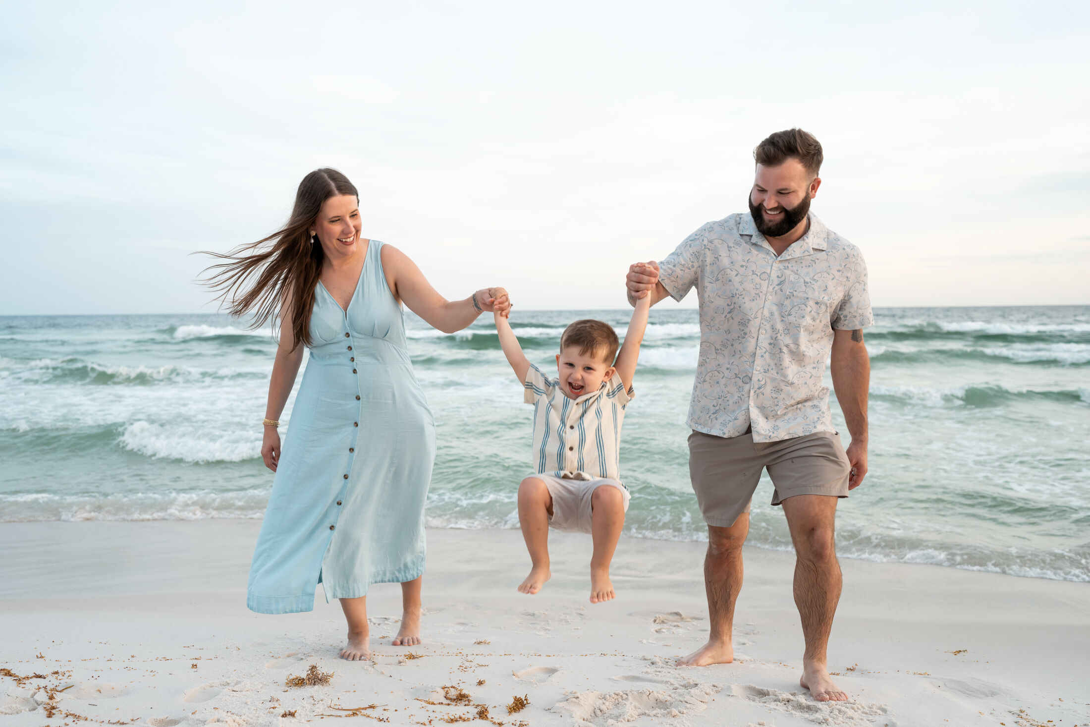 Parents swinging their laughing toddler between them at the shoreline of Navarre Beach at golden hour