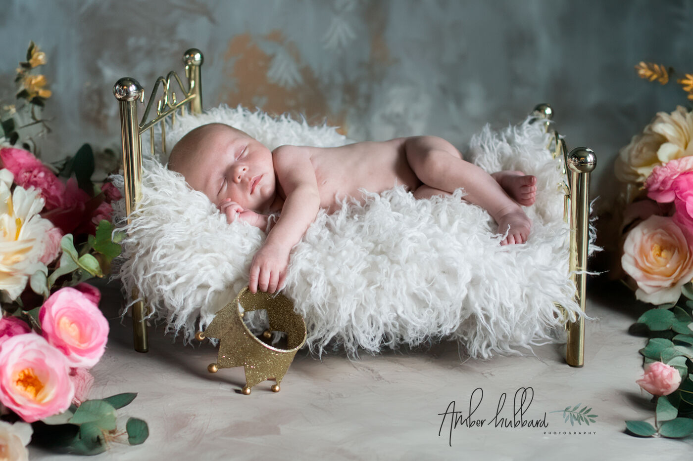 Newborn sleeping on a fluffy white blanket in a tiny brass bed surrounded by pink florals