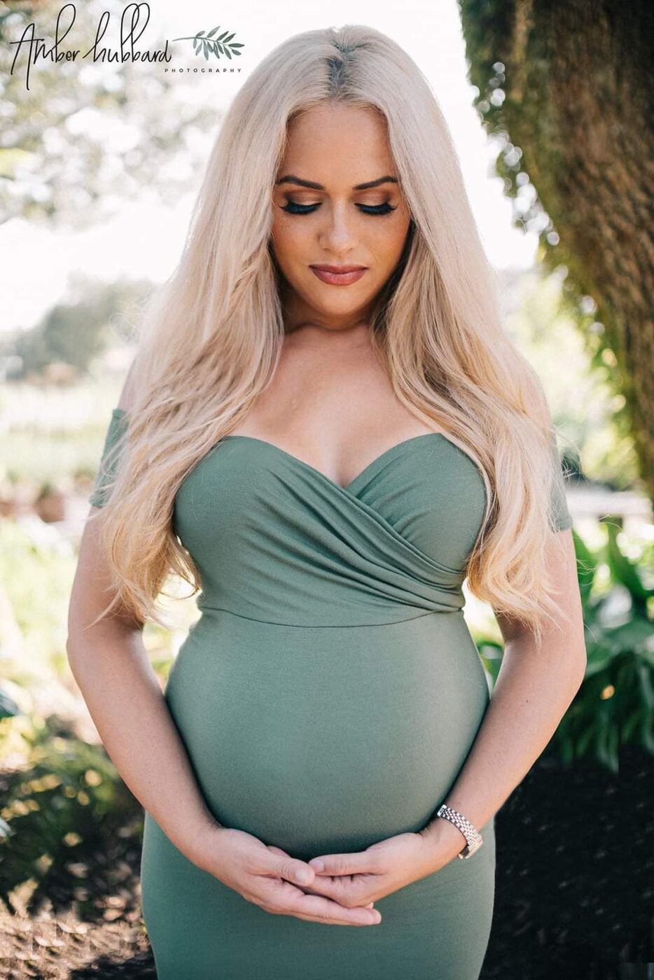 Expectant mother in a sage green dress cradling her belly beneath coastal trees